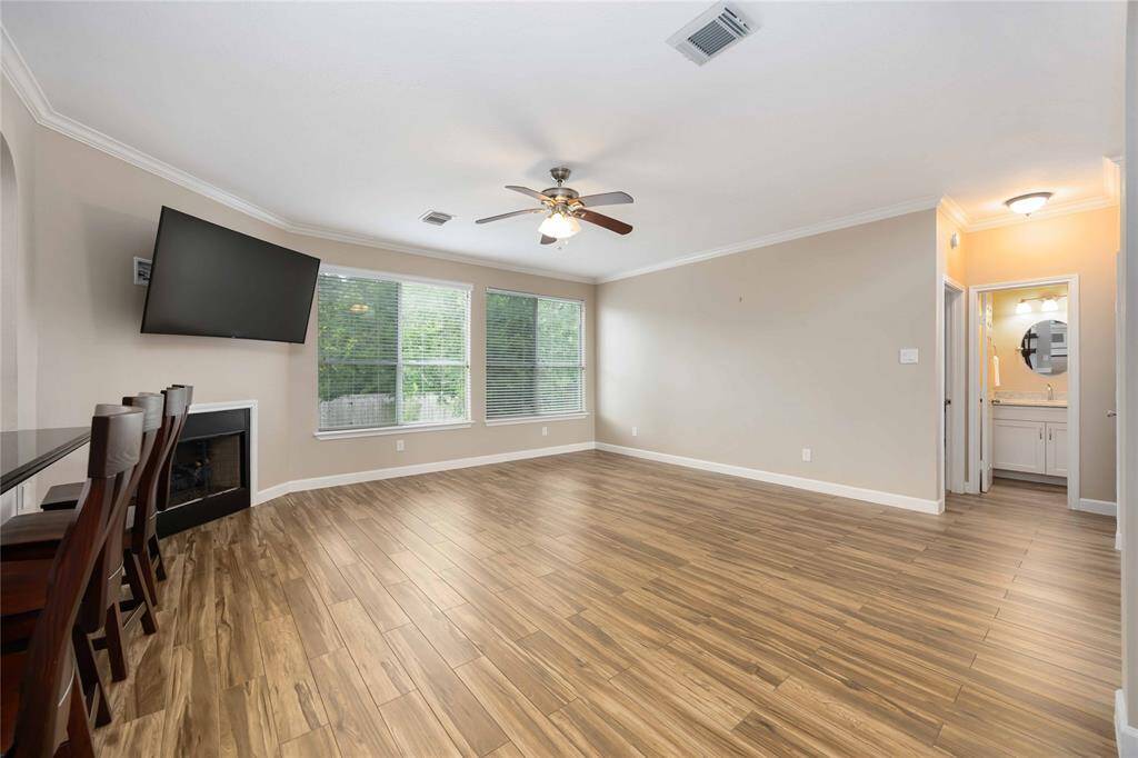 This spacious living room features wood-style flooring and a cozy fireplace. Large windows offer plenty of natural light, and there's a ceiling fan for added comfort. It connects to a kitchen area with bar seating and has access to a bright bathroom.