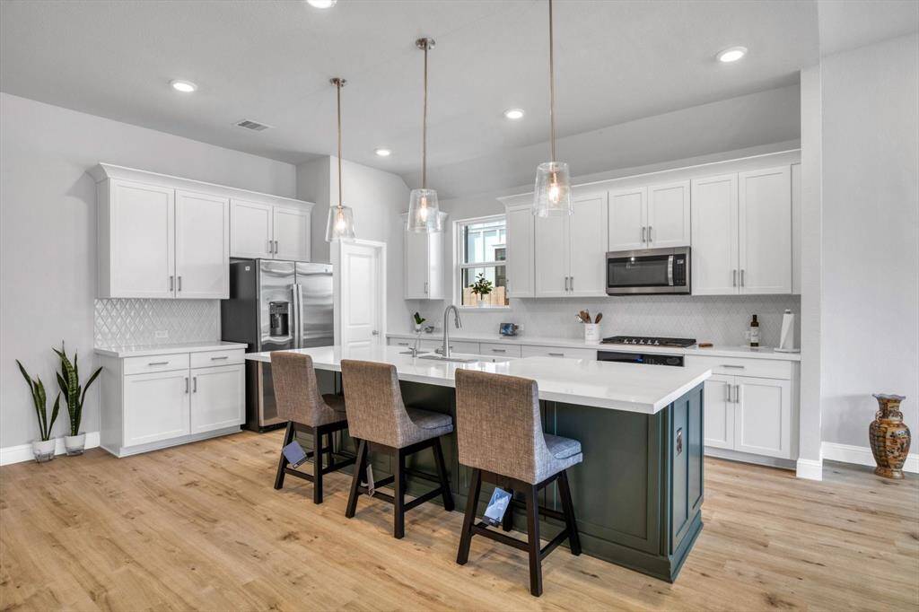 This modern kitchen features sleek white cabinetry, stainless steel appliances, and a spacious island with seating. The open layout and wood flooring add warmth, while pendant lighting provides a stylish touch.