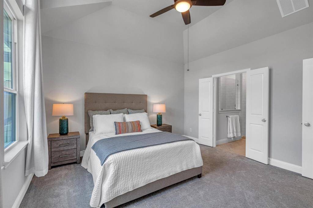 Primary bedroom with a vaulted ceiling and a ceiling fan, and an en-suite bathroom. Large windows provide plenty of natural light and a view of the backyard. The room includes a cozy bed, a dresser, and an open doorway leading to the living area with hardwood floors.
