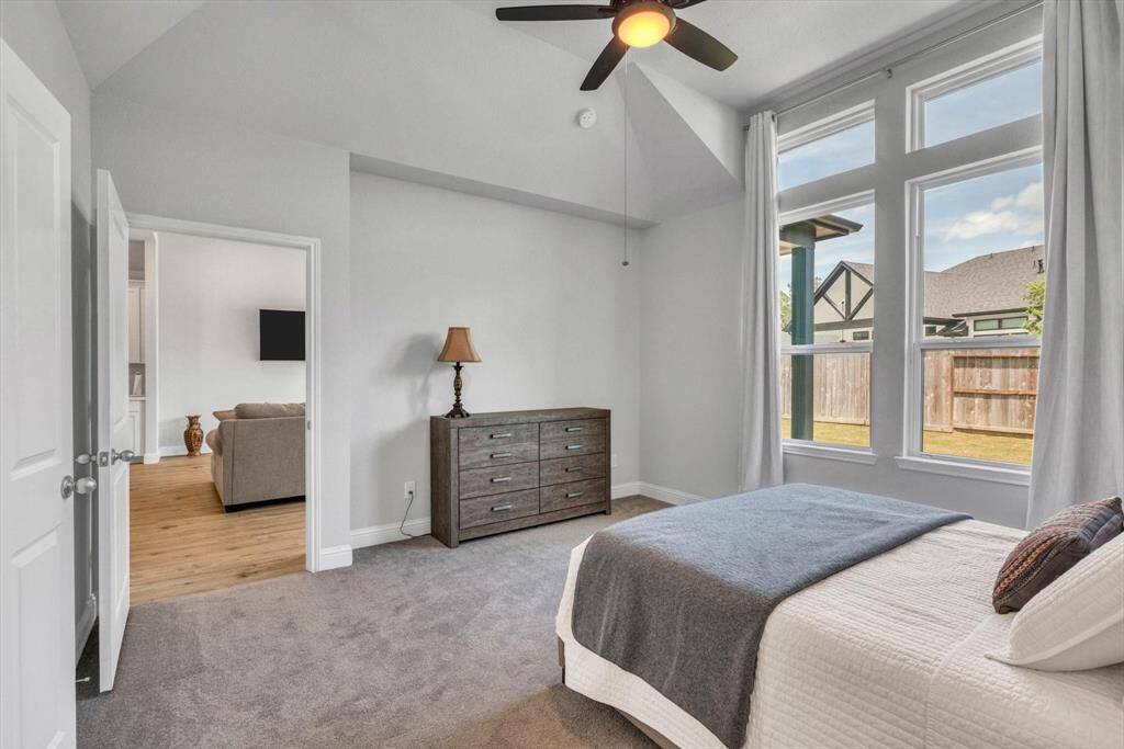 Primary bedroom with a vaulted ceiling and a ceiling fan, and an en-suite bathroom. Large windows provide plenty of natural light and a view of the backyard. The room includes a cozy bed, a dresser, and an open doorway leading to the living area with hardwood floors.