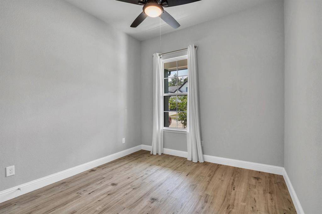 This room features light gray walls, wood-style flooring, and a ceiling fan. A window with white curtains allows natural light to fill the space, creating a bright and airy atmosphere.