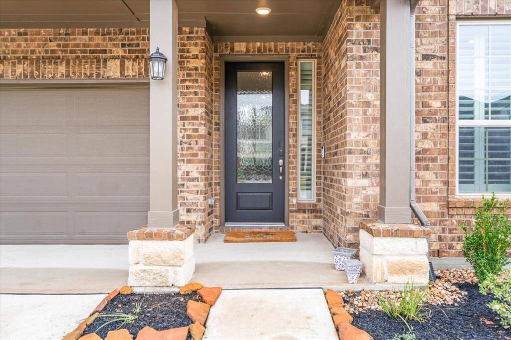 This photo showcases a welcoming brick facade with a covered entrance. The front door is modern with glass panels, flanked by a window covered in a Plantation shutter. The area features neatly landscaped elements and a two-car garage, offering a clean and inviting entryway to the home.
