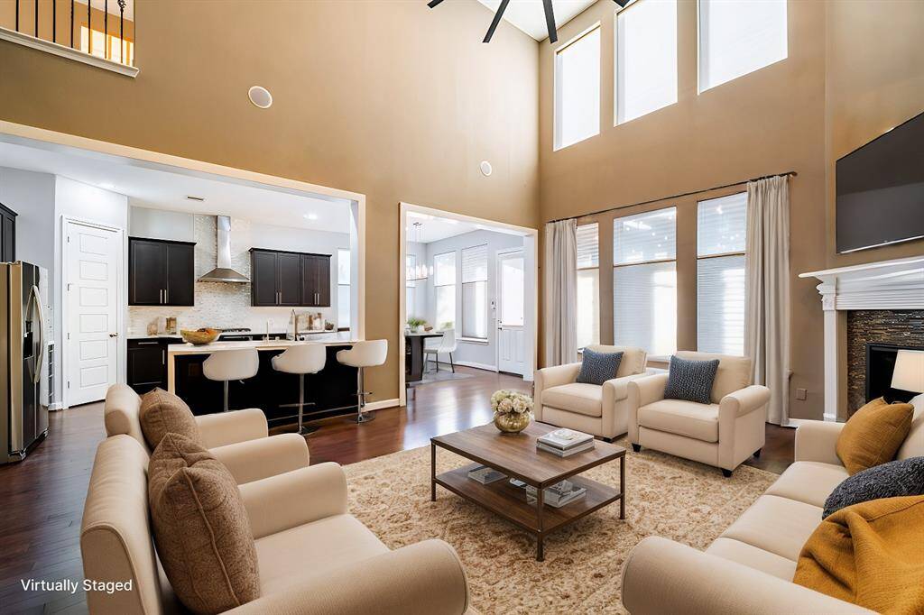 The open-concept space features soaring ceilings and expansive windows that fill the room with natural light. The living area flows seamlessly into a modern kitchen with dark cabinetry and a sleek island. A cozy fireplace adds warmth, while hardwood floors enhance the elegant design.