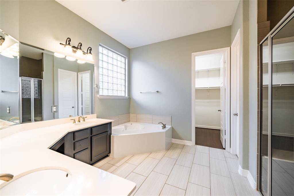 This luxurious primary bath offers a serene retreat with a jetted tub and a separate glass-enclosed shower. Dual vanities with ample counter space and stylish lighting provide both elegance and functionality. Annexed to the primary bedroom, it also features direct access to a spacious walk-in closet for ultimate convenience and organization. The large glass block window brings in natural light while maintaining privacy, completing this spa-like experience.