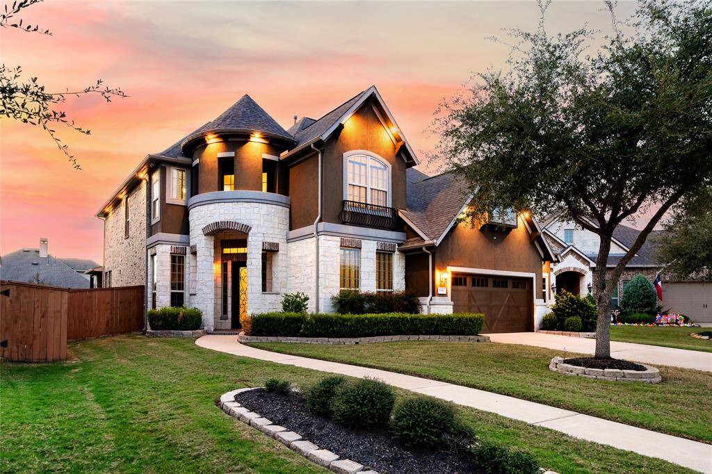 This stunning home showcases a mix of modern and classic design, beautifully illuminated by warm exterior lighting against a vibrant sunset backdrop. A neatly landscaped yard, bordered with shrubs and trees, enhances its curb appeal. The pitched roofline, Juliet balcony, and spacious two-car garage complete the perfect blend of charm and functionality.