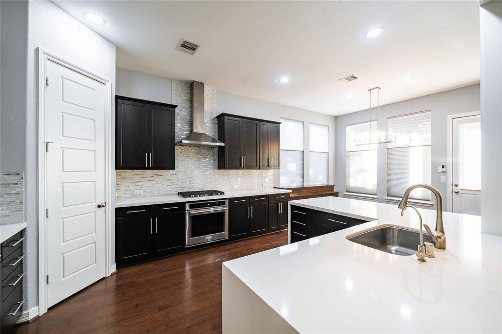 This bright and airy kitchen features a functional layout with ample natural light streaming through large windows. A built-in gas cooktop with a sleek stainless-steel hood is centered on a contemporary tile backsplash, adding both style and practicality. The spacious island with an undermount sink and extended quartz countertops provides generous prep and serving space. A modern chandelier accents the dining nook, blending seamlessly with the minimalist design for a refined yet welcoming vibe.