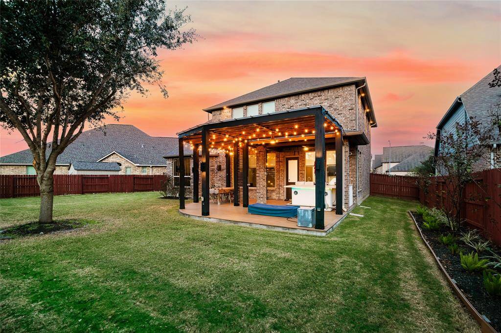 This spacious backyard is perfect for outdoor living, featuring a stunning pergola adorned with string lights that create a warm, inviting ambiance. The covered patio offers ample space for dining and entertaining, complete with a built-in countertop for added convenience. The lush green lawn is bordered by a wooden fence for privacy, while the vibrant landscaping adds natural beauty. The sunset backdrop enhances the charm, making this outdoor space ideal for relaxation or hosting gatherings.