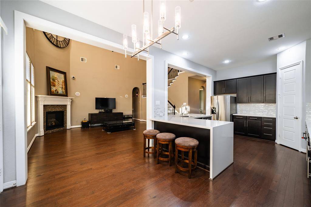 This open-concept space seamlessly connects the kitchen, dining, and living areas, perfect for modern living. The kitchen features sleek dark cabinets, a spacious island with a waterfall quartz countertop, and stylish pendant lighting. The living area boasts soaring ceilings, a cozy fireplace with a detailed mantel, and ample natural light from large windows. Hardwood floors run throughout, enhancing the home's elegance.