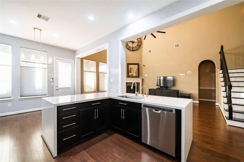 This inviting kitchen features a modern U-shaped layout with sleek black cabinetry and polished quartz countertops. The stainless-steel sink and dishwasher are seamlessly integrated into the island, which overlooks the spacious living area with high ceilings, a fireplace, and elegant décor. Large windows allow ample natural light, while the adjacent dining area is accented with a stylish chandelier. The open staircase and arched entryway enhance the home’s airy and cohesive design.