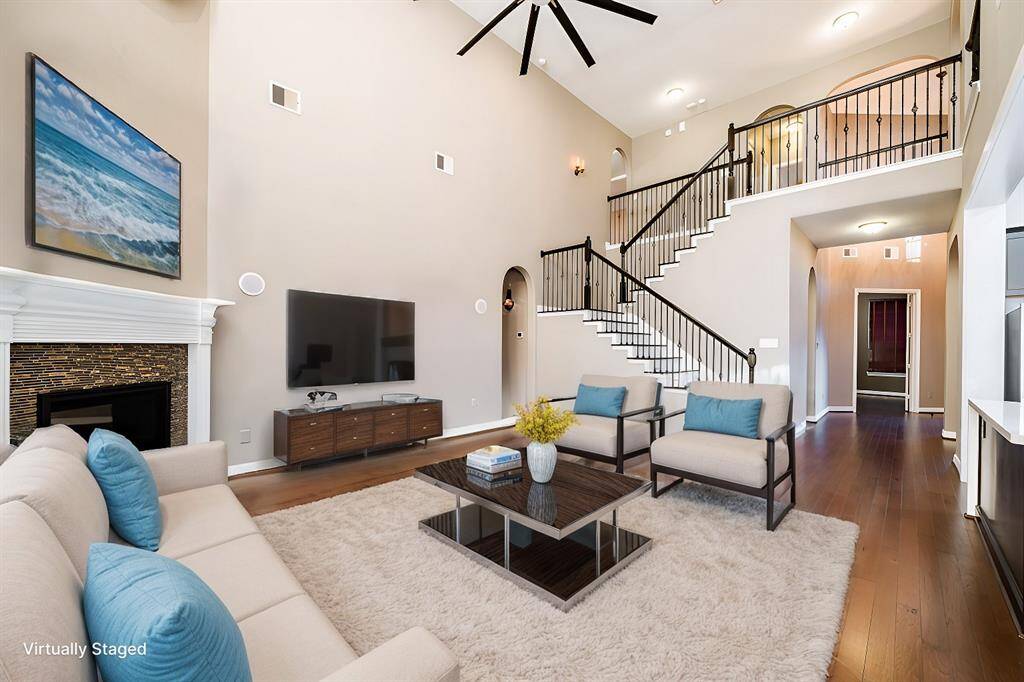 Living room features soaring ceilings and warm hardwood floors, creating a spacious and inviting ambiance. A sleek fireplace with a detailed mantel serves as the focal point, complemented by wrought iron staircase railings. Natural light enhances the open layout, ideal for relaxation or entertaining.