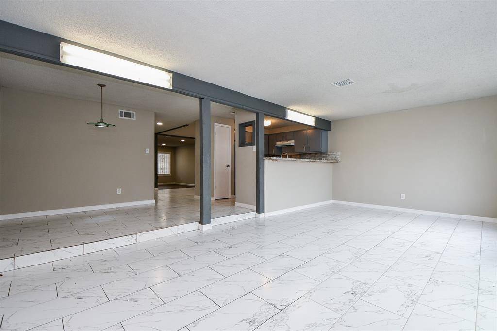 Spacious room with light-colored tile flooring and neutral walls, featuring an open layout that connects to a kitchen with a breakfast bar. Recessed lighting and a large window provide ample natural light.