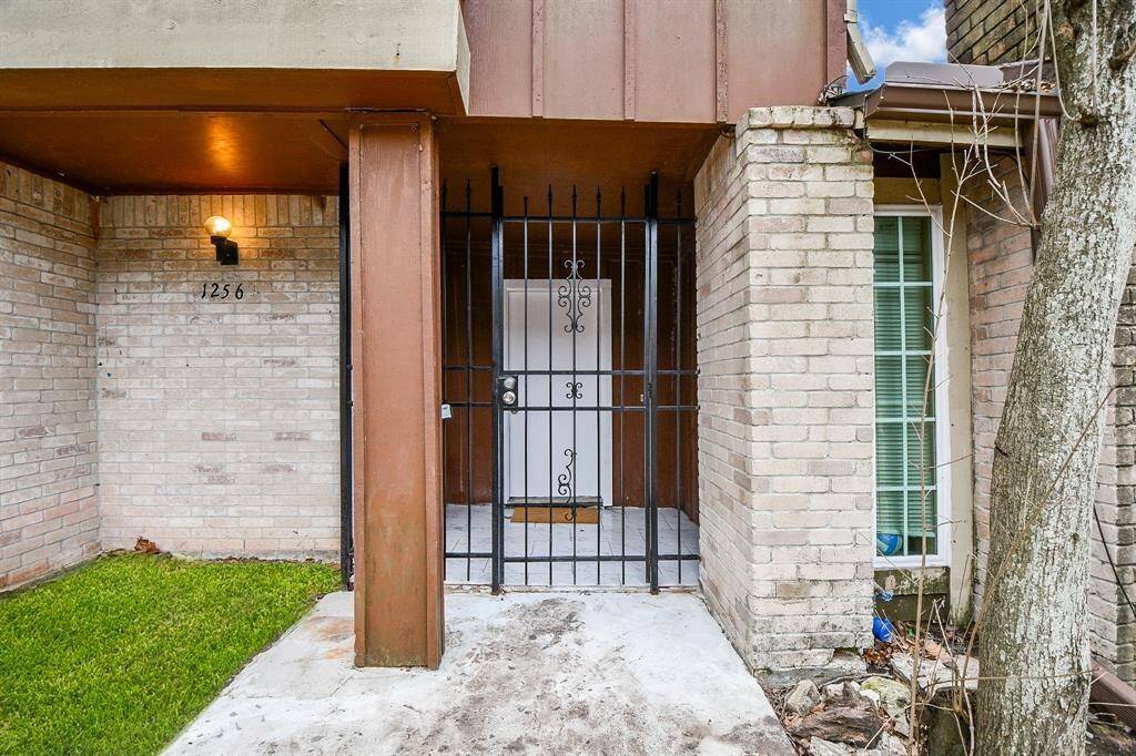 This photo shows the entrance of a home with a gated entryway. It features brick walls, a small grassy area to the left, and a window to the right. A light fixture is mounted above the house number.