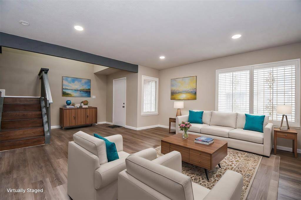 This photo showcases a bright, modern living room with hardwood floors, stylish furniture, and large windows. The neutral walls complement the vibrant decor accents. A staircase adds architectural interest, and the space feels open and inviting. Virtual staging was used.