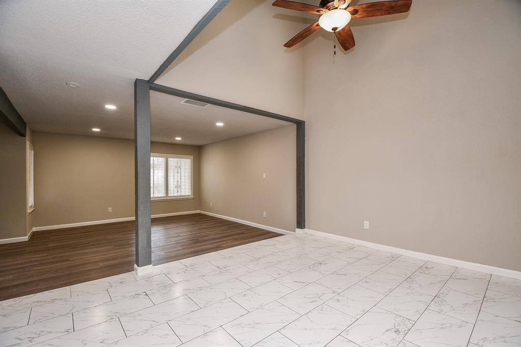 This spacious room features a vaulted ceiling with a ceiling fan, a mix of wood and tile flooring, and neutral walls. The area is divided by a beam, offering distinct spaces with ample natural light from a large window.