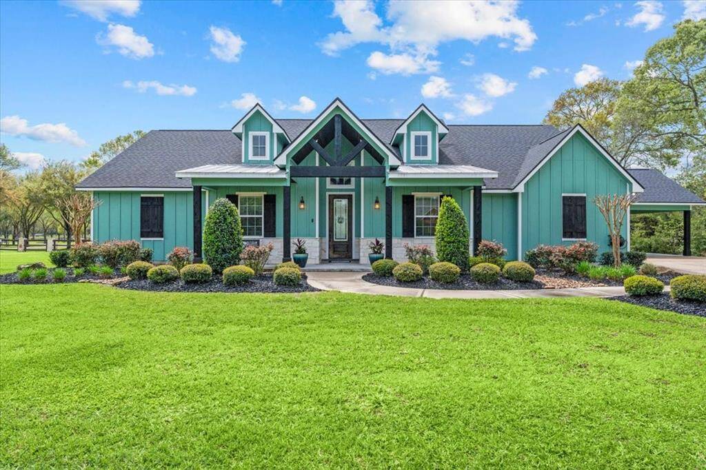 This charming single-story custom home features a vibrant exterior with a welcoming front porch. Surrounded by manicured landscaping and lush greenery, it offers a serene country feel with modern touches. The driveway leads to a convenient oversized garage carport on the side.