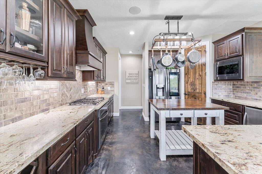 This kitchen features rich dark wood cabinets, granite countertops, gas cooktop, and a central island with a rustic finish. The space is accented by a pot rack, stainless steel appliances, wine glass holder, large pantry and a stylish backsplash, creating a warm and functional cooking area.