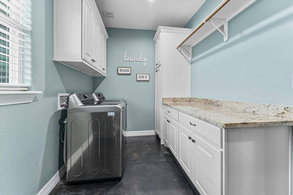 This room showcases a spacious laundry room with a washer and dryer, ample cabinetry, and granite countertop for all your folding needs. The space is bright, with light walls and a window bringing in natural light. There’s a hanging rod for your clean clothes too! This laundry room has enough room for a deep freeze or extra refrigerator.