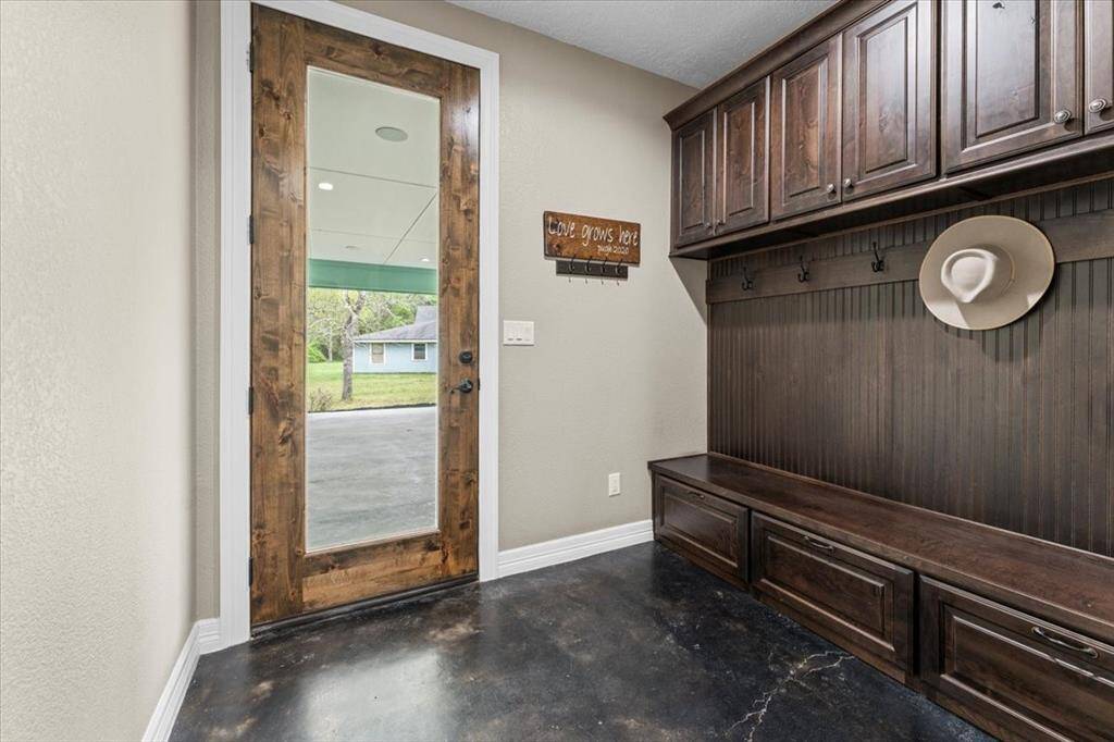 This photo shows a well-appointed mudroom with dark wood cabinetry, a bench with storage, coat hooks, and a glass door leading outside to the carport. It has a modern, rustic feel with polished concrete flooring.