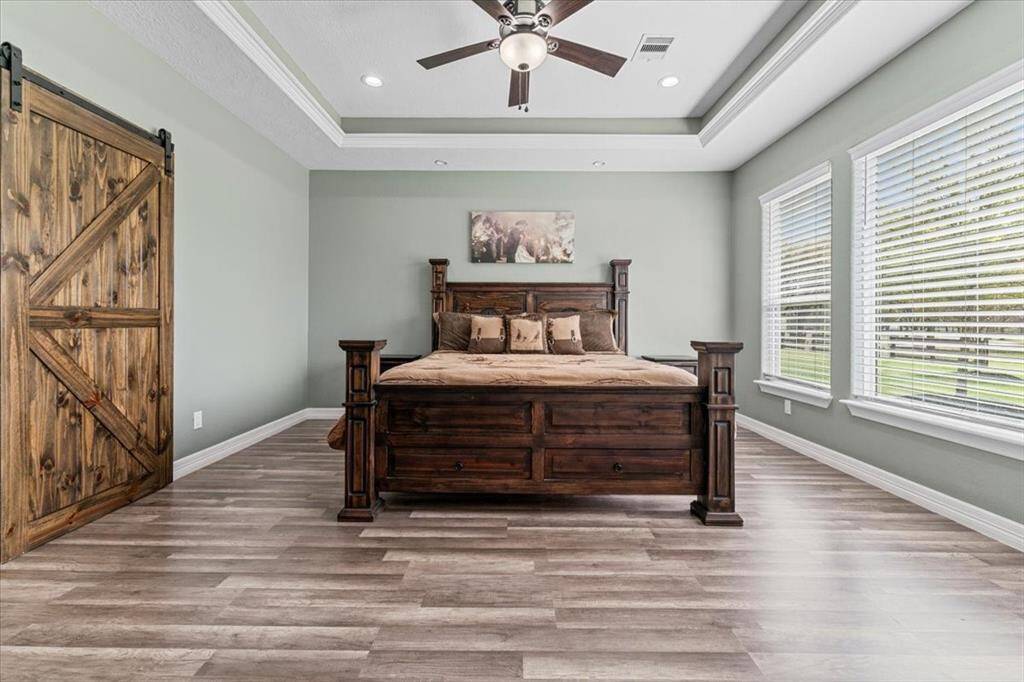 This spacious bedroom features a rustic wooden bed, sliding barn door, and large windows with abundant natural light. The room has neutral flooring, soft painted walls, and a ceiling fan for comfort.