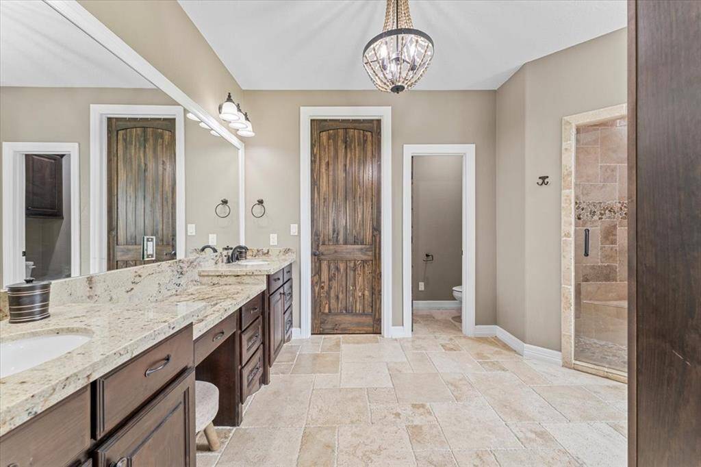 This spacious bathroom features elegant beige tile flooring, large granite countertop with dual sinks, a vanity area and dark wood cabinetry. It includes a separate walk-in shower with stone accents and a separate toilet area. This bathroom comes with a soaking tub for you to relax at the end of a long day. Its stylish chandelier provides elegance and matching wall sconces provide ample lighting, complementing the rustic wooden doors.