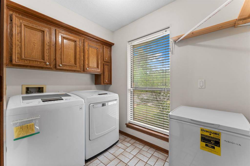 This laundry room is bright and functional, featuring a washer and dryer set, upper wooden cabinets for storage, a large window providing natural light, and a compact freezer.