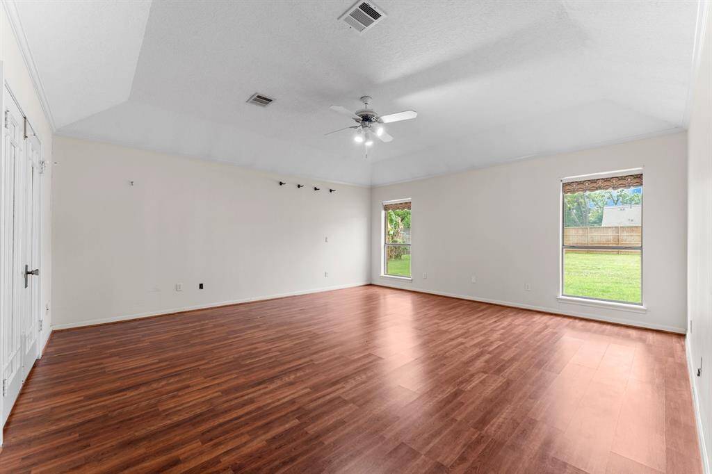 This spacious room features warm wood flooring, neutral walls, a ceiling fan, and ample natural light from two large windows overlooking a fenced backyard. Perfect for a living area or versatile space.