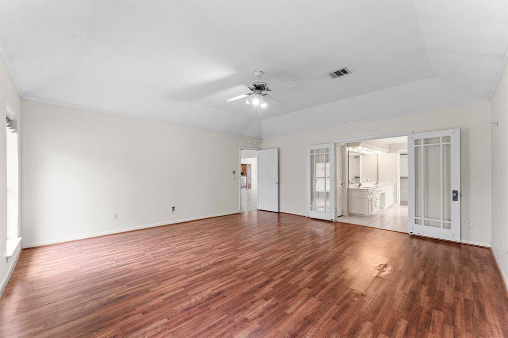 This spacious room features hardwood floors, neutral walls, and a ceiling fan. It has ample natural light and sliding glass doors leading to a bright bathroom with dual sinks.