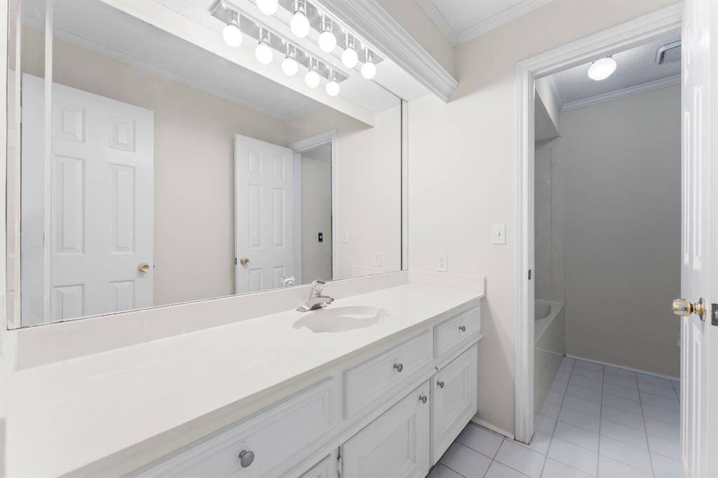This bathroom features a large mirror above a spacious white countertop with ample cabinetry below. It has bright lighting and a door leading to a shower area, with clean tile flooring.