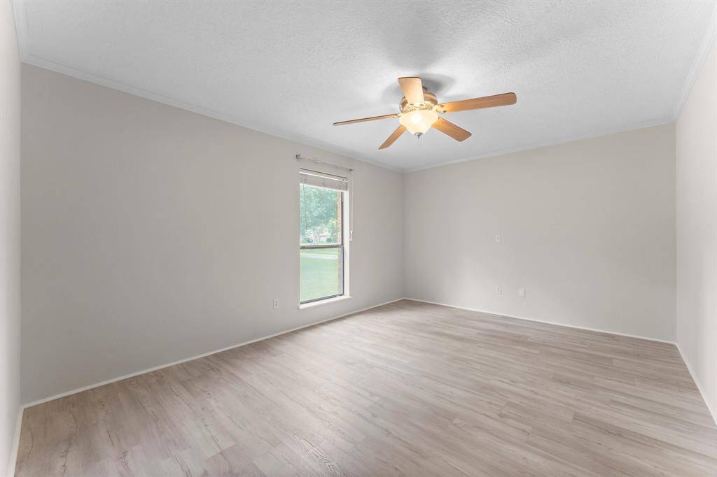 Bright, spacious room with light wood flooring, neutral walls, a ceiling fan, and a large window offering a view of greenery.