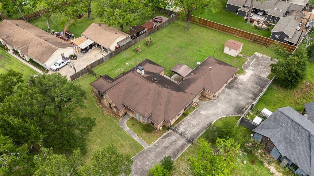 This aerial view showcases a spacious single-story home with a brown roof and a large driveway. The property features a sizable backyard, mature trees, and is situated in a quiet neighborhood with nearby homes.