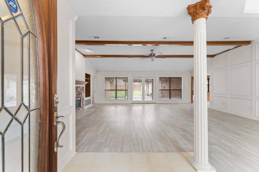 This spacious open-concept living area features elegant wood beams, a classic fireplace, and abundant natural light from large windows and glass doors leading to the backyard. The room has light wood flooring and decorative columns, creating an inviting and airy atmosphere.