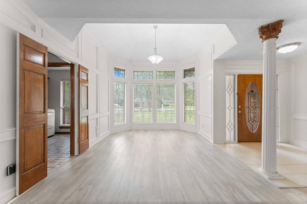This spacious entryway features elegant stained glass windows, a warm wooden door, and light hardwood flooring. The area is bright and welcoming, enhanced by decorative columns and classic paneling, offering a sophisticated first impression.