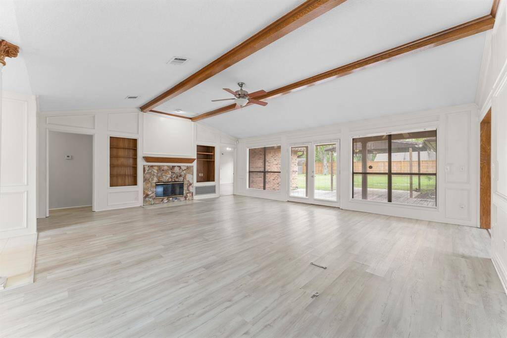 This spacious living room features vaulted ceilings with wooden beams, a stone fireplace, built-in shelving, and large windows offering plenty of natural light and views of the backyard.