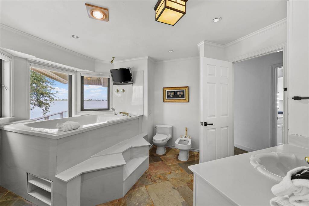 Indulge in a spa-like retreat with a raised soaking tub framed by windows offering waterfront views and mounted TV for the ultimate in luxury comfort.