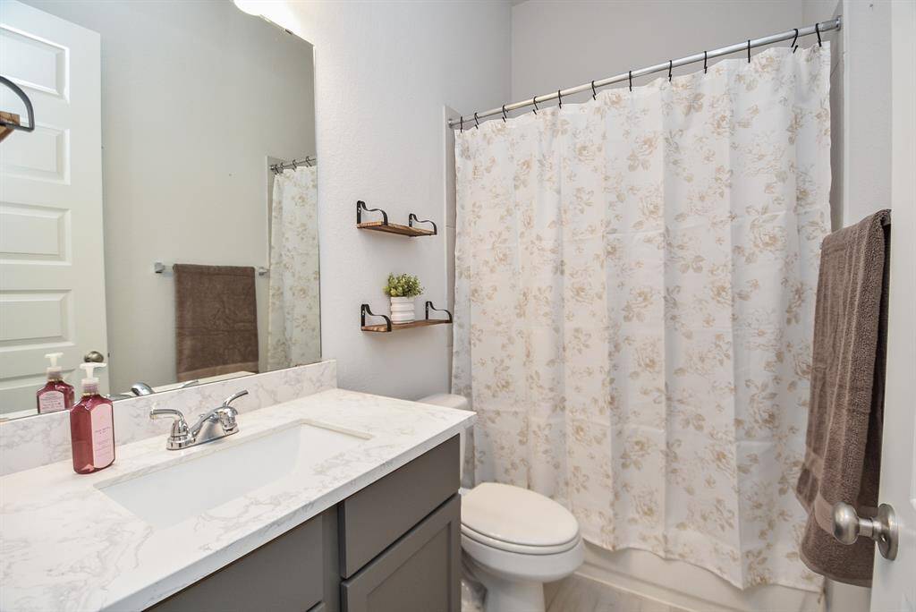 This is a modern bathroom featuring a sleek vanity with a marble countertop, a large mirror, and a combination bathtub/shower with a floral curtain. Neutral tones and small decorative shelves add a cozy touch.