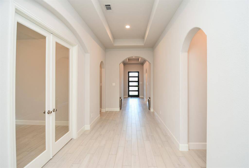 A bright, spacious hallway features elegant archways and recessed lighting. Double glass doors on the left lead to an adjacent room, and a modern glass-paneled front door is visible at the end.