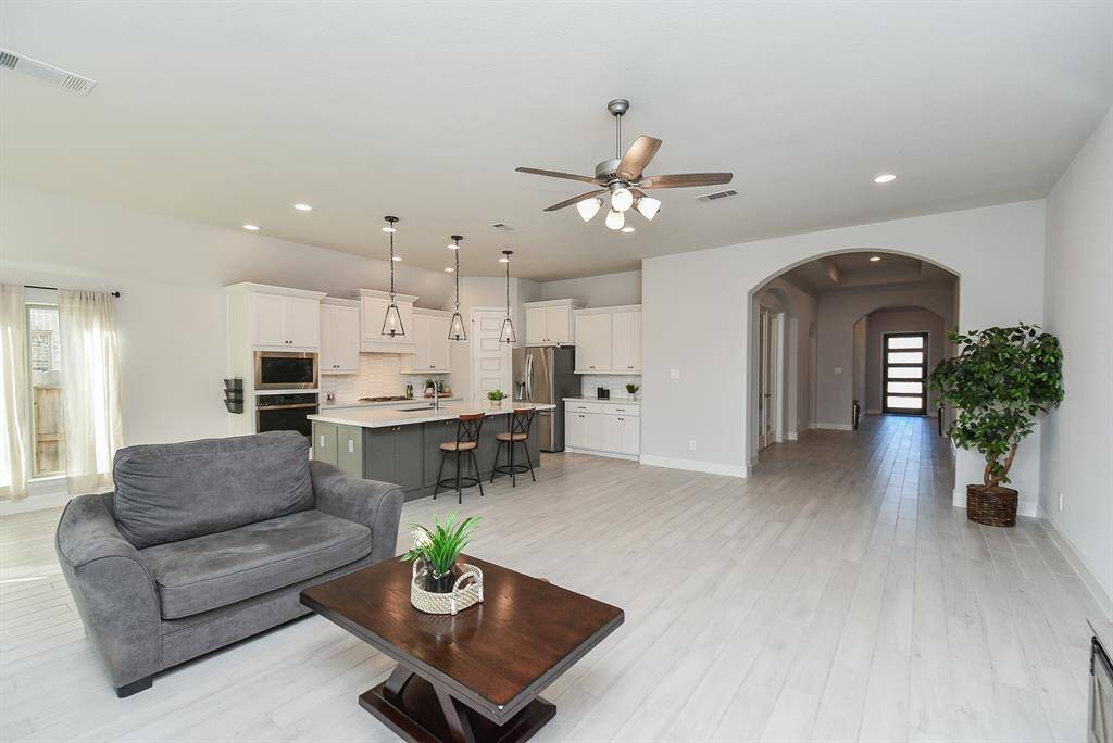 This photo shows a spacious open-concept living area with a modern kitchen with white cabinetry and island seating, and a cozy living space. A ceiling fan and pendant lights add style, while an arched hallway leads to other rooms. Natural light filters through sheer curtains from a patio door.