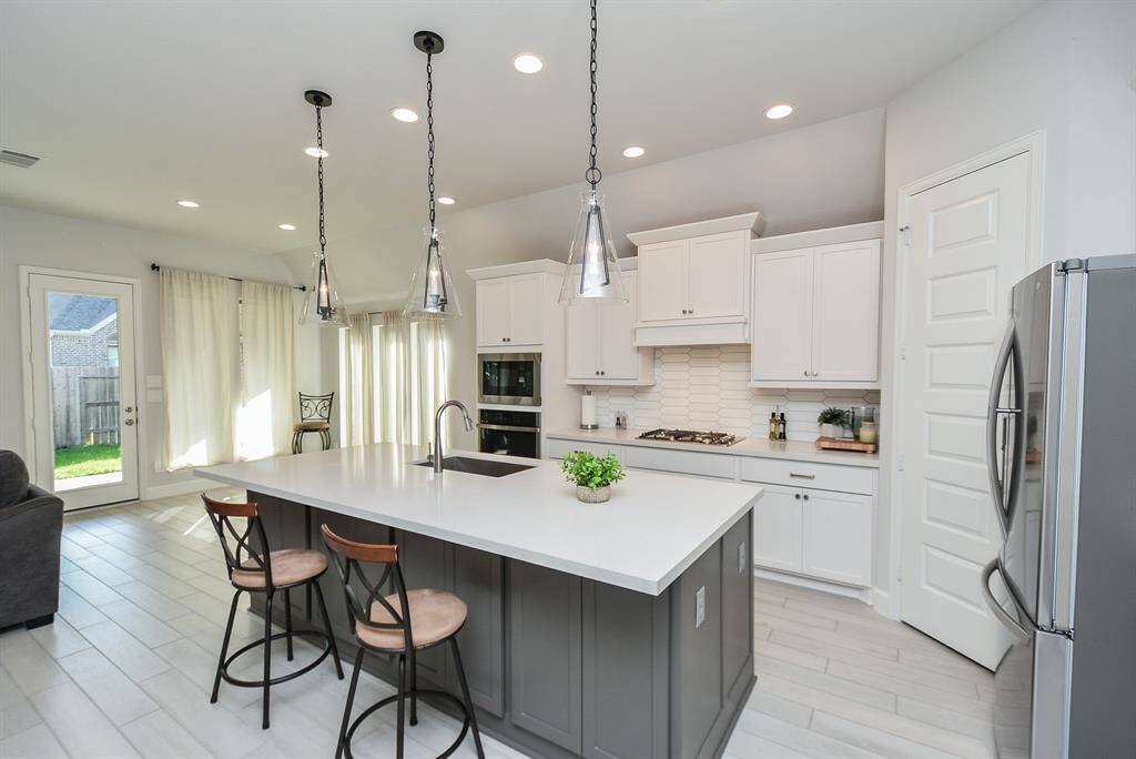 This modern kitchen features a spacious island with seating, sleek pendant lighting, and white cabinetry. It includes stainless steel appliances, a built-in oven, and a bright, open layout with large windows and a door leading to a backyard.