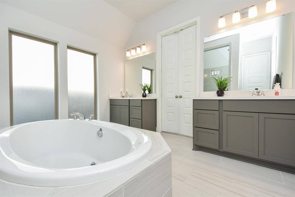 This bathroom features a spacious soaking tub, dual vanities with modern fixtures, and ample natural light from large frosted windows. The sleek cabinetry adds a contemporary touch.