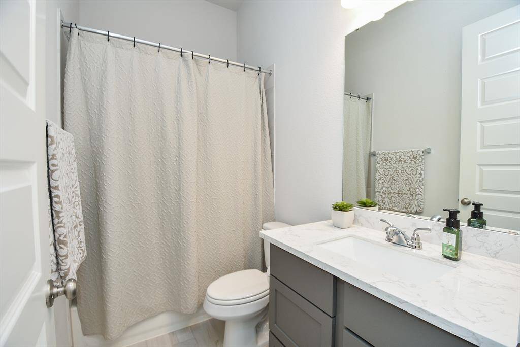 This bathroom features a modern design with a sleek white countertop, under-mount sink, and gray cabinetry. It includes a shower with a curtain, a large mirror, and subtle decorative touches for a clean, inviting look.