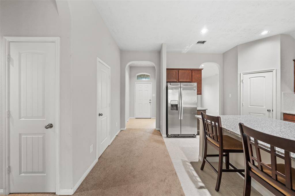 This photo showcases a modern kitchen and hallway with neutral walls and carpet. It features a stainless steel refrigerator, granite countertops, and a dining bar with seating. The space is bright with recessed lighting and has multiple doorways leading to other rooms.