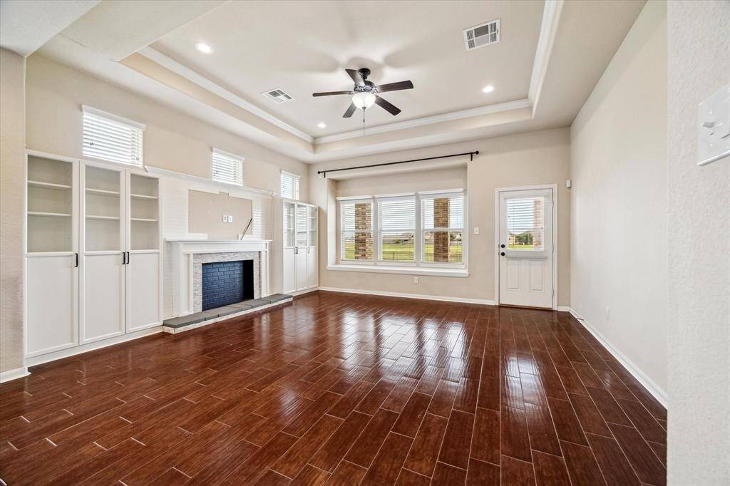 This spacious family room offers a warm and inviting atmosphere with rich wood-look tile flooring, a tray ceiling with crown molding, and recessed lighting. Built-in cabinets flank a charming mock fireplace, while a wall of windows and a glass door showcase serene backyard views and fill the room with natural light.