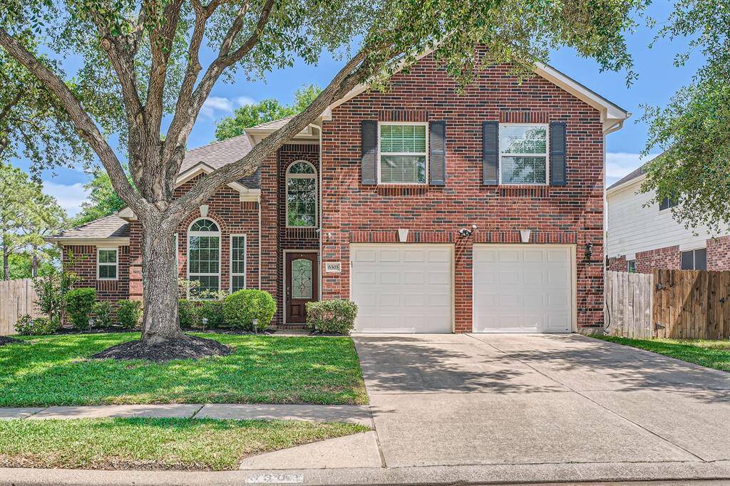This charming home features a classic red brick exterior with a welcoming arched entryway and large front windows. It has a spacious two-car garage and is beautifully framed by mature trees and well-maintained landscaping, offering great curb appeal.