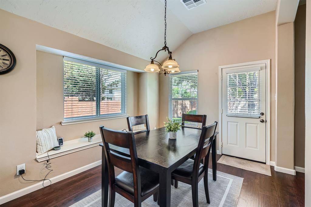 This photo shows a bright dining area with a high ceiling, featuring a dark wood table and chairs. It has large windows with blinds, allowing natural light to fill the space. A small bench sits beneath the window, and there's a door leading outside, enhancing the room's accessibility and charm.
