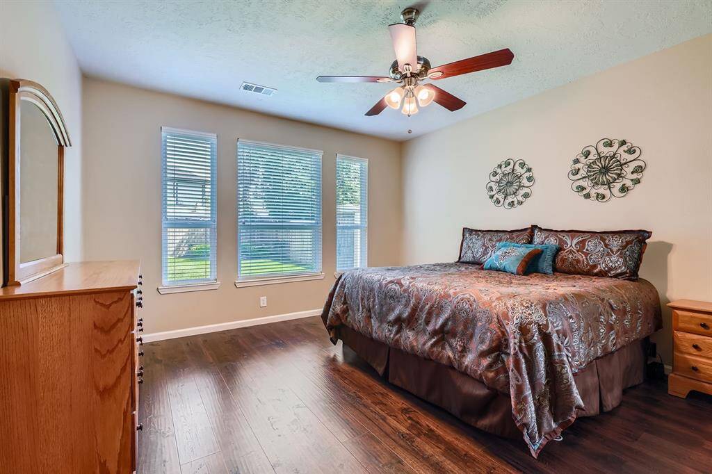 This bedroom features large windows with natural light, wood floors, and a ceiling fan. It has a cozy atmosphere with neutral walls and decorative accents, offering ample space for relaxation.