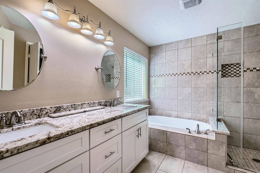 This bathroom features a modern design with a dual-sink granite countertop, round mirrors, and stylish lighting. It includes a tiled shower and a separate bathtub, offering a clean and elegant space.