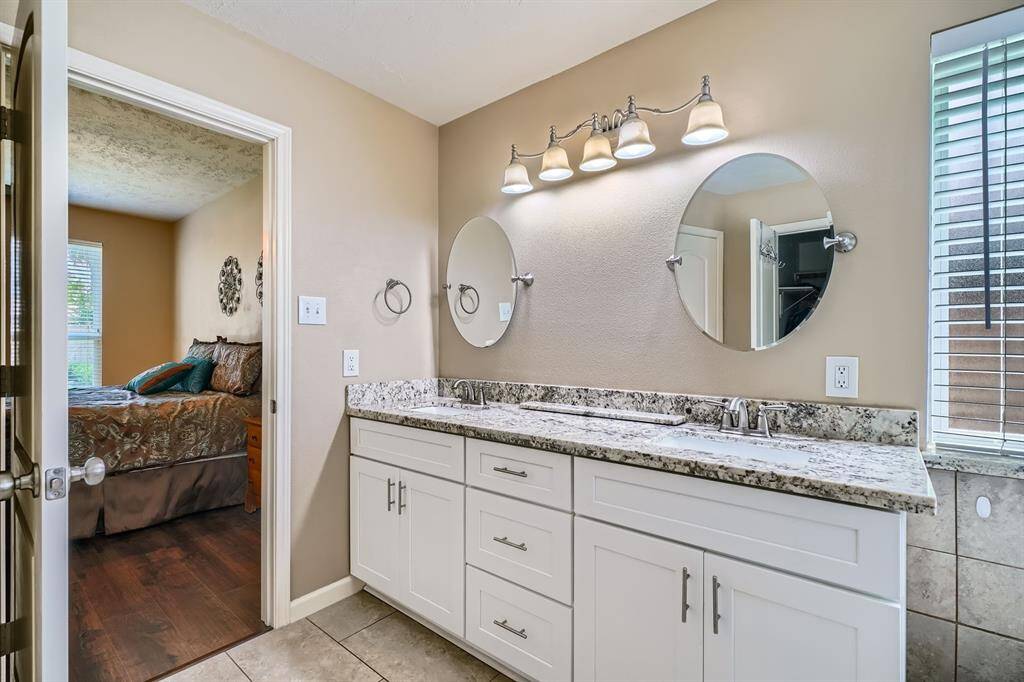 This photo features a modern bathroom with dual sinks, granite countertops, and circular mirrors. It connects to a cozy bedroom, visible through an open door. Natural light filters through a window with blinds.