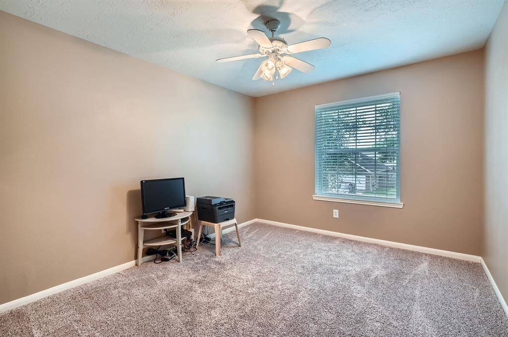 This room features a neutral color scheme with beige walls and carpet flooring, a ceiling fan, and a window providing natural light. It's currently set up as a small office with a desk and computer.