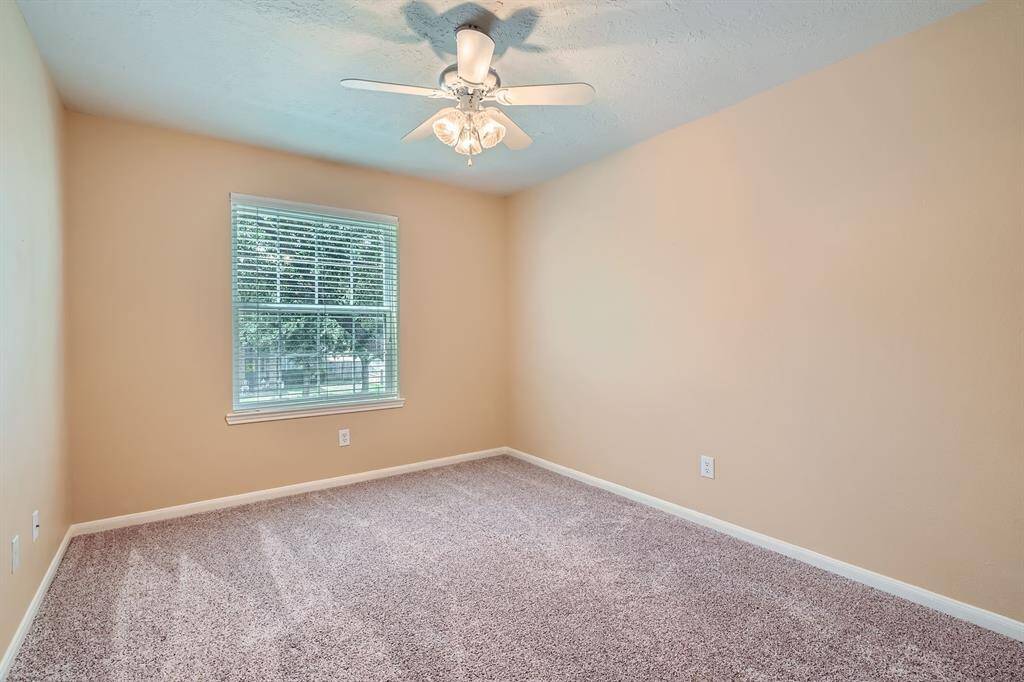 This room features soft beige walls and a plush carpet, offering a cozy atmosphere. A ceiling fan and a large window provide light and ventilation, perfect for a comfortable living space.