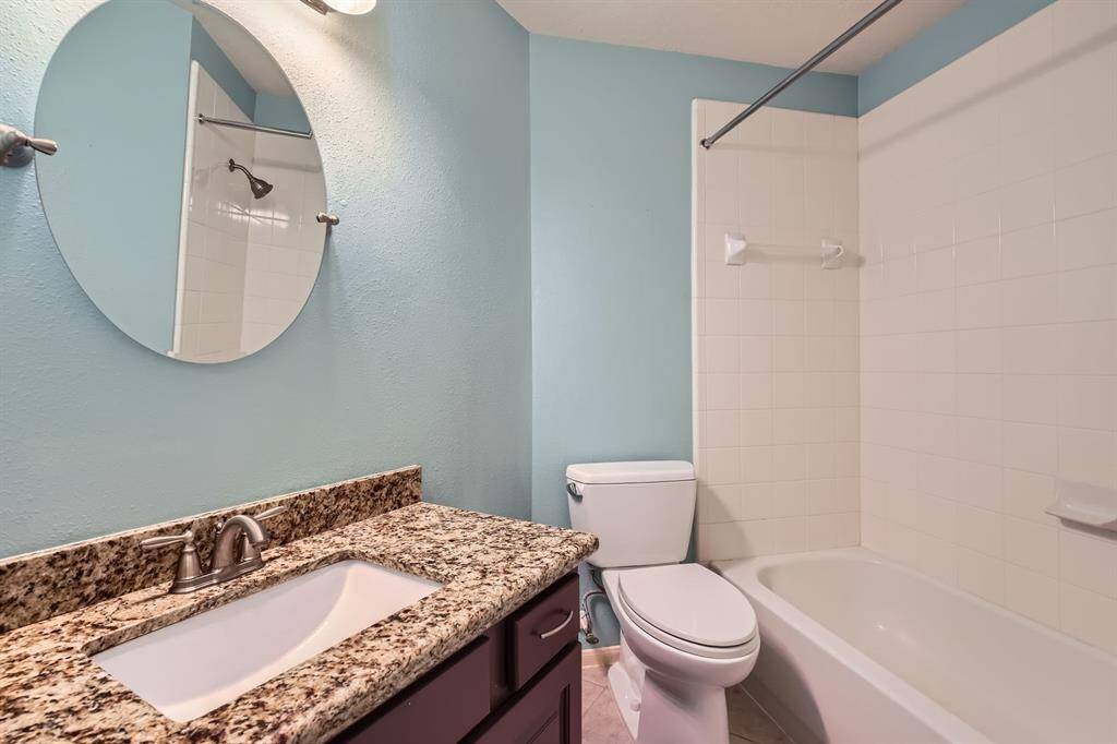 This bathroom features a clean, modern design with a granite countertop, under-mount sink, and a sleek round mirror. The space is accentuated by soft blue walls, a white bathtub with a shower, and contemporary fixtures.
